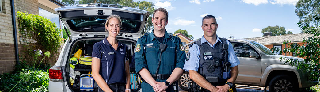 A uniformed police officer, a uniformed paramedic and a mental health clinician standing together in front of a car