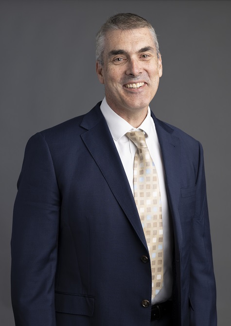 A portrait photo of Executive General Manager Corporate Lachlan Funnell with cropped short grey hair, a smile on his face and a navy suit with white shirt and silver, gold and white tie. 